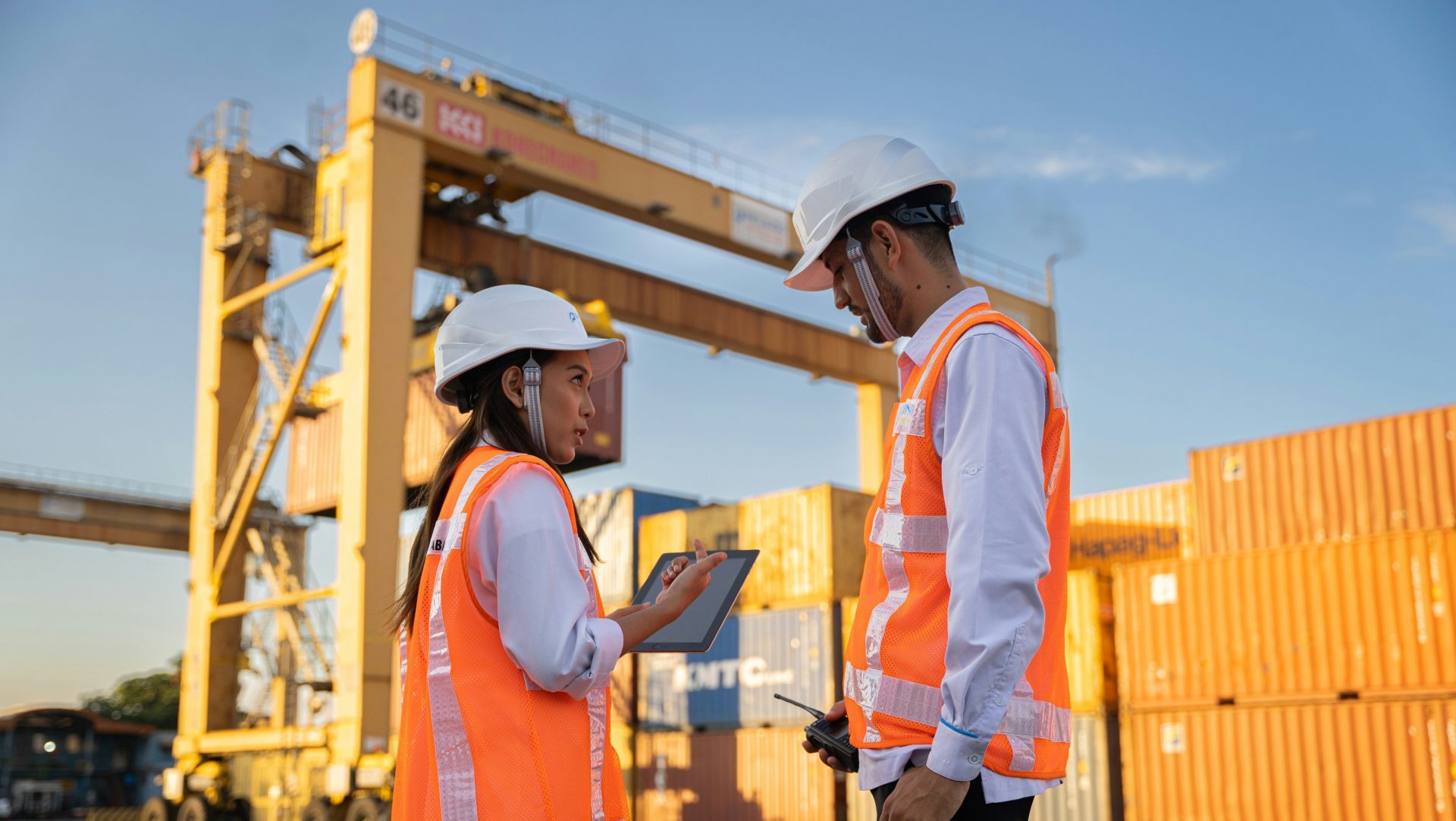 Two workers in hard hats discuss plans near shipping containers.